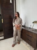 Person wearing a beige Leinen outfit with sunglasses and sandals, standing next to a wooden dresser.